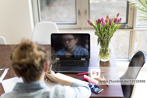 Rückansicht einer Frau mit Laptop-Computer  die zu Hause auf einem Buch schreibt