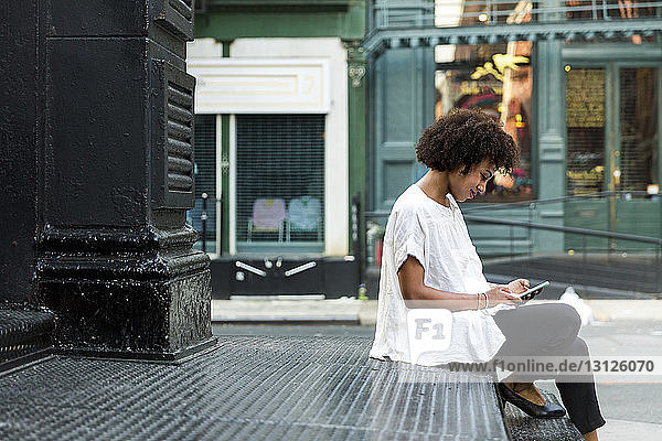 Seitenansicht einer Frau  die ein Smartphone benutzt  während sie auf einer Treppe in der Stadt sitzt