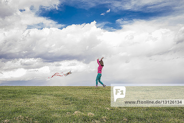 Mädchen fliegt Drachen in voller Länge auf Grasfeld vor bewölktem Himmel