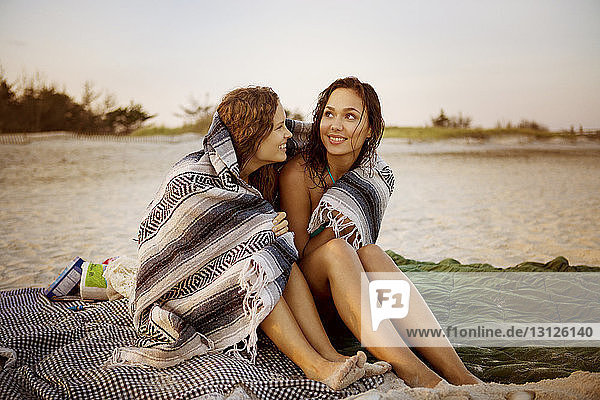 Lächelnde Frauen in eine Decke gehüllt am Strand sitzend