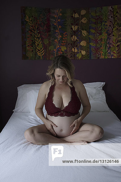 Schwangere Frau in Dessous berührt den Bauch  während sie zu Hause auf dem Bett sitzt