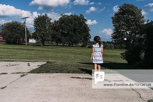 Rear view of girl standing on footpath by grassy field at park during sunny day