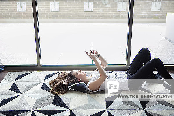 Junge Frau benutzt Smartphone  während sie zu Hause am Fenster auf dem Teppich liegt