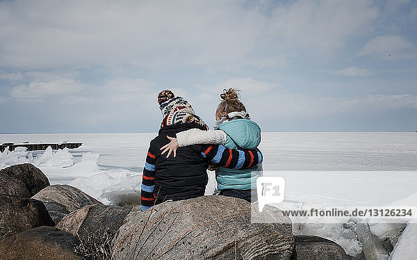 Rear view of siblings sitting on rock by frozen lake against cloudy sky
