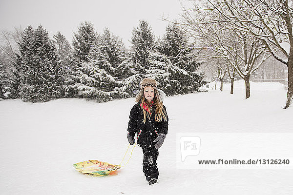 Full length of girl wearing warm clothing while walking with sled on snowy field against trees during snowfall