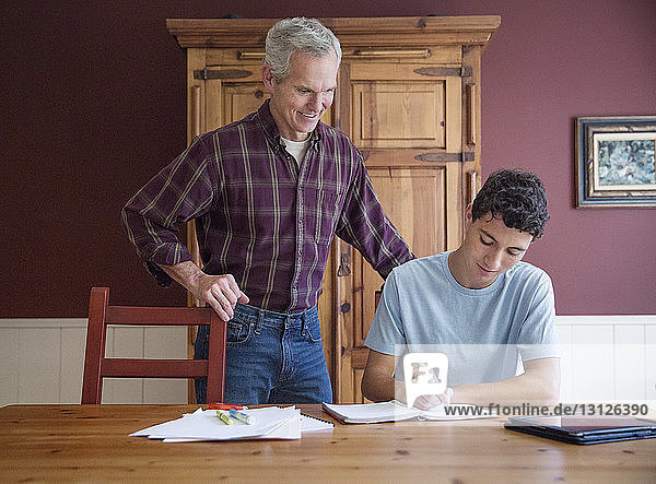 Lächelnder älterer Mann betrachtet Enkel  der zu Hause am Tisch lernt
