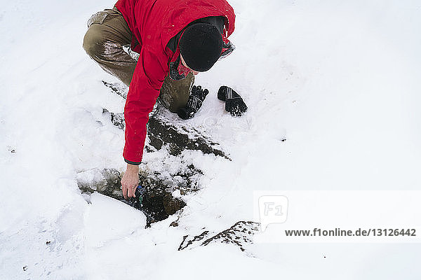 High angle view of hiker filling water bottle from stream during winter