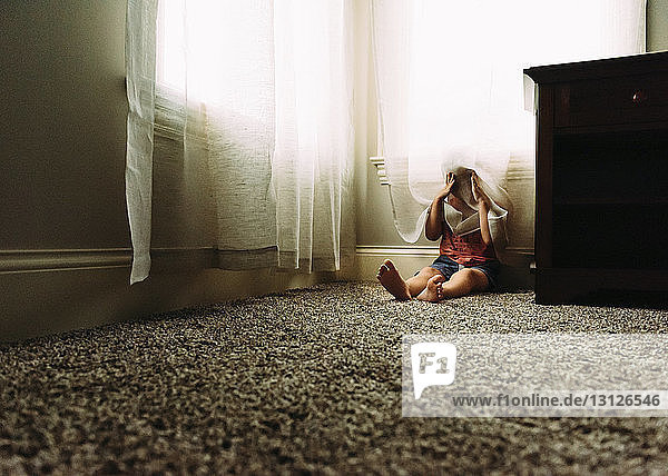Verspielter Junge  der sich hinter einem Vorhang versteckt  während er zu Hause auf dem Teppich sitzt