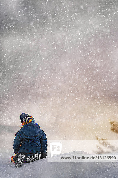 Rear view of boy sitting on snow covered field during snowfall at sunset