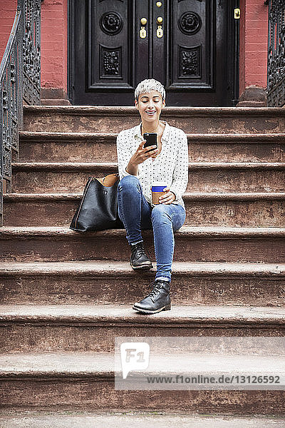 Glückliche Frau benutzt Mobiltelefon  während sie mit Kaffeetasse auf der Treppe sitzt