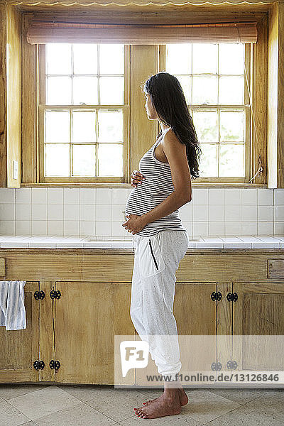 Nachdenkliche schwangere Frau berührt Bauch  während sie in der Küche steht