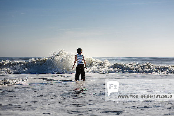 Rückansicht eines Jungen  der mit Wellen spielt  während er bei Sonnenuntergang im Meer gegen den Himmel steht