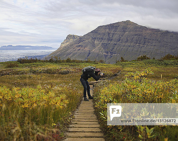 Seitenansicht eines Wanderers mit Rucksack  der mit der Kamera fotografiert  während er sich auf dem Feld gegen Berge beugt