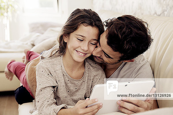 Father embracing daughter with digital tablet while resting on sofa
