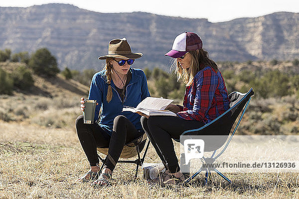 Freundinnen schauen sich ein Buch an  während sie bei Sonnenschein auf Campingstühlen auf einem Feld gegen die Berge sitzen