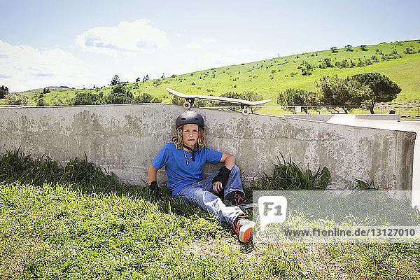 Portrait of confident boy sitting on field at skateboard park