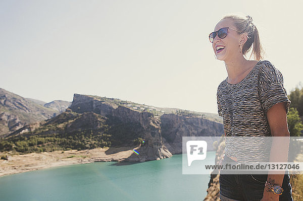 Glückliche Frau mit Sonnenbrille schaut weg  während sie gegen klaren Himmel und Fluss steht