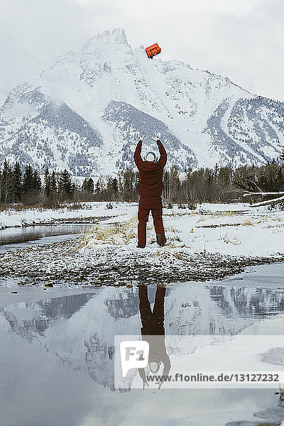 Rear view of man throwing box in air against snowcapped mountains while standing by lake