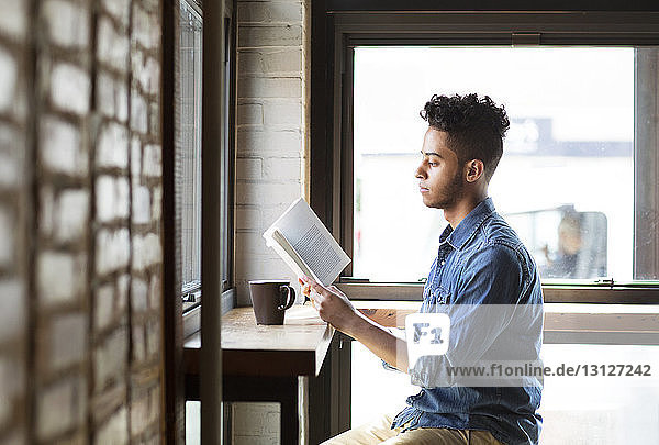 Seitenansicht eines Mannes  der ein Buch liest  während er an den Fenstern eines Cafés sitzt