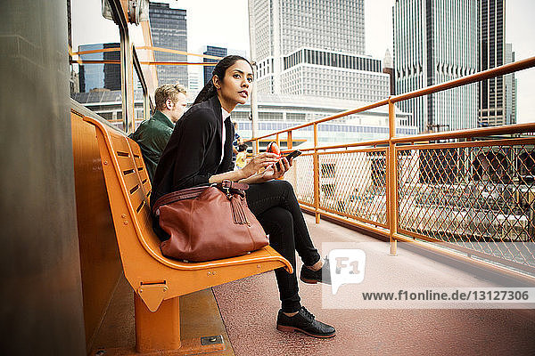 Thoughtful woman using phone while sitting on bench in city