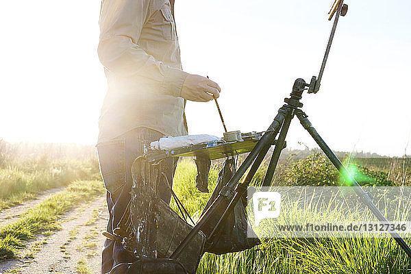 Midsection of painter standing by easel at field during sunny day
