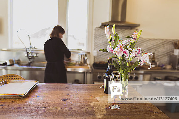 Weinglas neben Vase auf Tisch mit Frau  die zu Hause im Hintergrund arbeitet