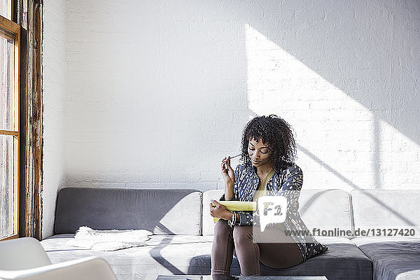 Junge Geschäftsfrau liest Buch  während sie im Kreativbüro auf dem Sofa sitzt