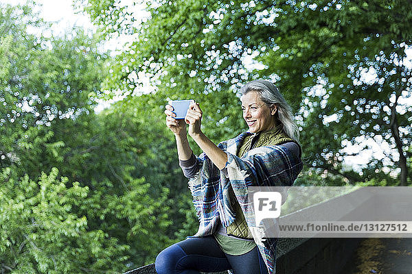 Woman photographing through smart phone while standing on footpath against branches