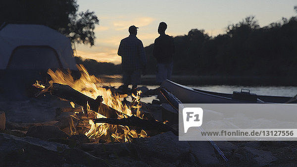 Silhouette Vater und Sohn am Seeufer stehend mit brennendem Lagerfeuer im Vordergrund