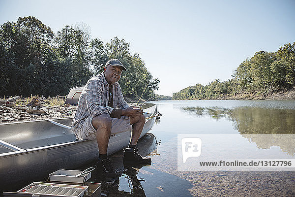 Portrait of man holding fishing rod while sitting on boat at lakeshore