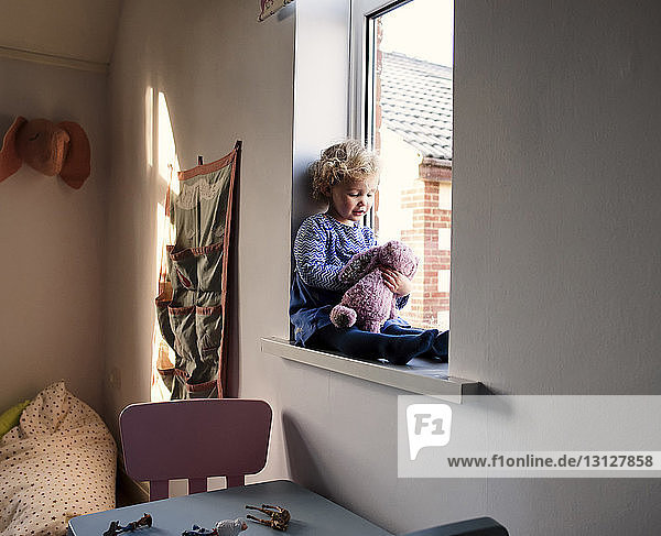 Mädchen spielt mit Stofftier  während sie zu Hause auf dem Fensterbrett sitzt