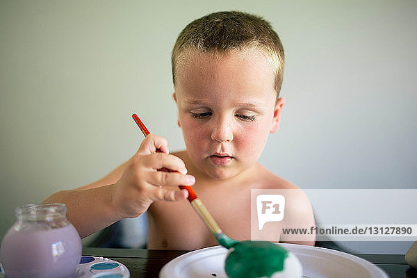 Junge ohne Hemd färbt Ei  während er zu Hause am Tisch sitzt