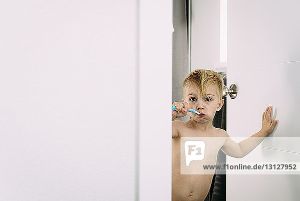 Portrait of shirtless boy brushing teeth while standing at doorway in bathroom