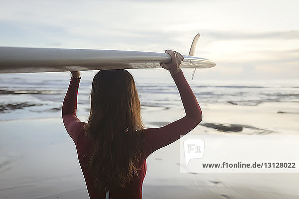 Rückansicht einer jungen Frau  die am Strand stehend ein Surfbrett auf dem Kopf trägt