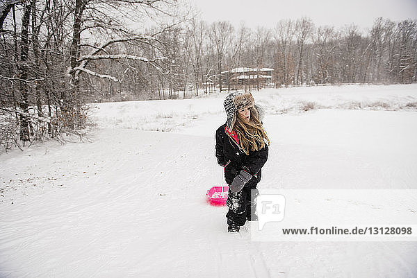 Girl wearing warm clothing while walking with sled on snowy field against trees during snowfall