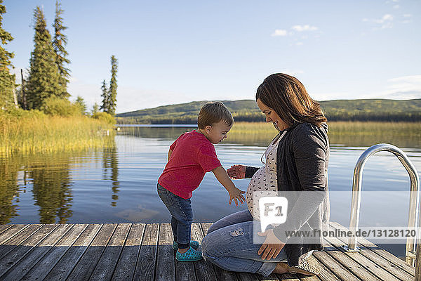 Side view of son touching pregnant mother's belly on pier over lake