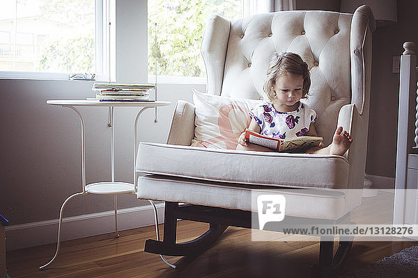 Mädchen liest Bilderbuch  während sie zu Hause auf einem Schaukelstuhl sitzt