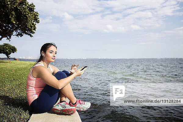 Thoughtful woman holding smart phone and sitting by sea against cloudy sky