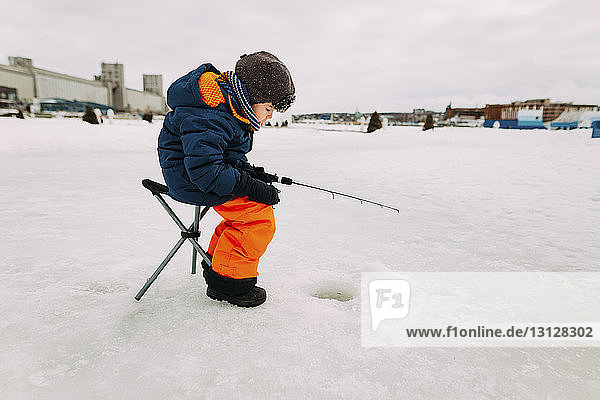 Side view of boy fishing in frozen lake while sitting on stool against sky