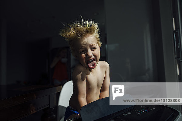 Porträt eines Jungen ohne Hemd  der die Zunge herausstreckt  während er die Brise der Klimaanlage zu Hause genießt