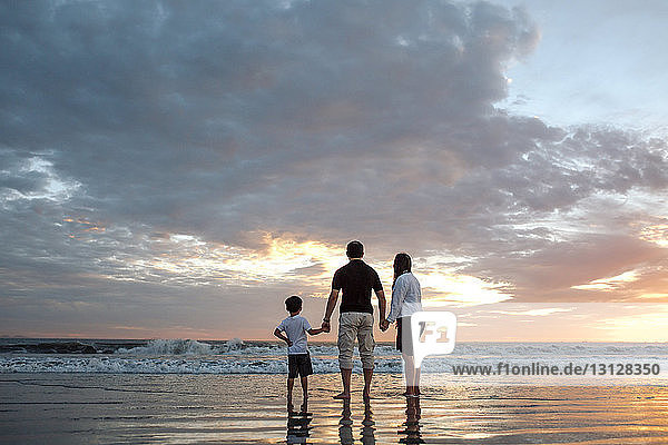 Rückansicht einer Familie  die bei Sonnenuntergang am Strand vor bewölktem Himmel Händchen hält