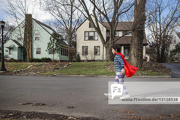 Seitenansicht eines Jungen mit rotem Umhang  der auf der Straße gegen kahle Bäume und Häuser läuft