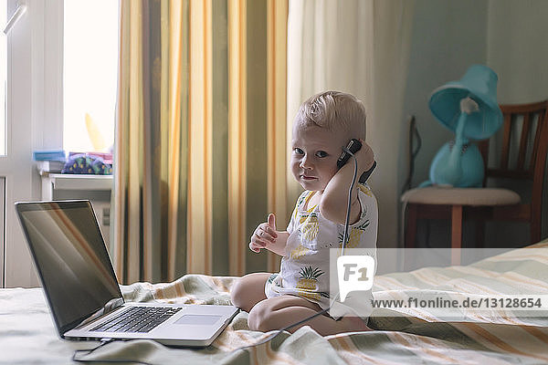 Porträt eines süßen kleinen Jungen mit Mobiltelefon und Laptop-Computer auf dem Bett zu Hause