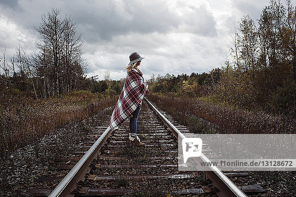 Side view of woman with blanket standing on railroad track against stormy clouds