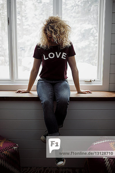 Frau in voller Länge trägt ein T-Shirt mit dem Text LOVE  während sie zu Hause auf dem Fensterbrett sitzt