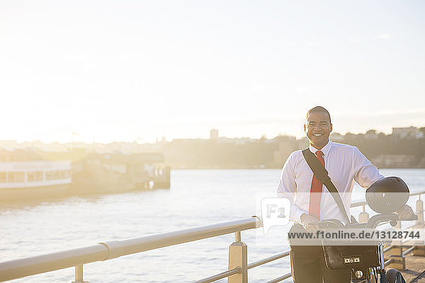 Portrait of smiling businessman with bicycle standing against river