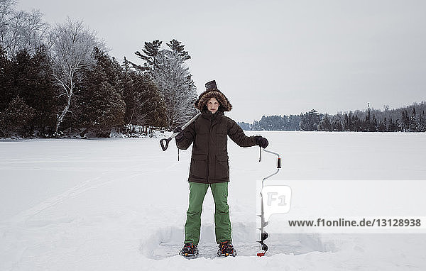 Porträt eines Mannes  der auf einem zugefrorenen See stehend Eisbohrer und Schaufel gegen den Himmel hält