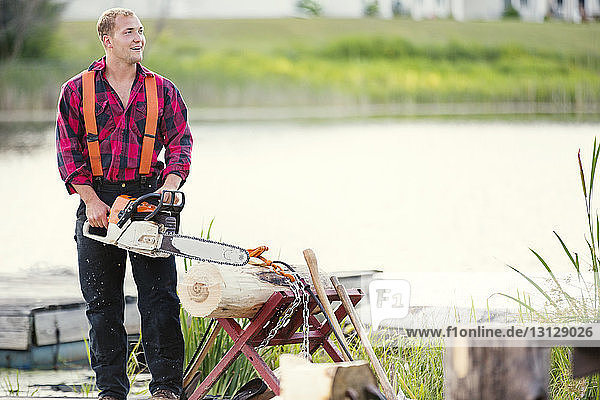 Smiling man with chainsaw cutting log at riverbank