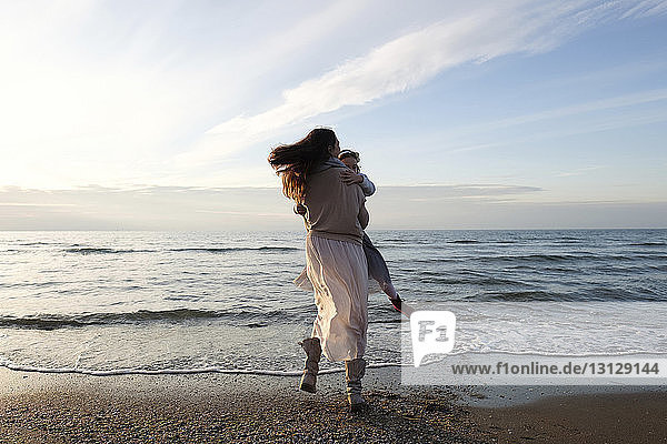 Rückansicht einer glücklichen Mutter  die bei Sonnenuntergang ihre Tochter am Strand gegen den Himmel dreht