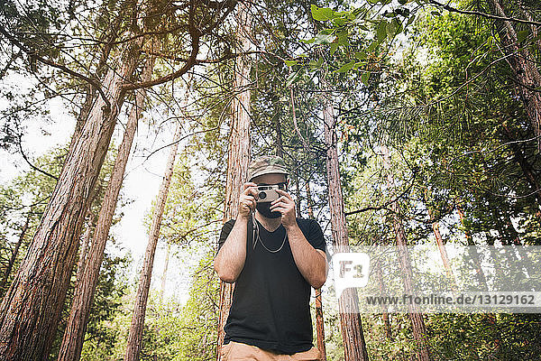 Man photographing through camera while standing against trees in forest